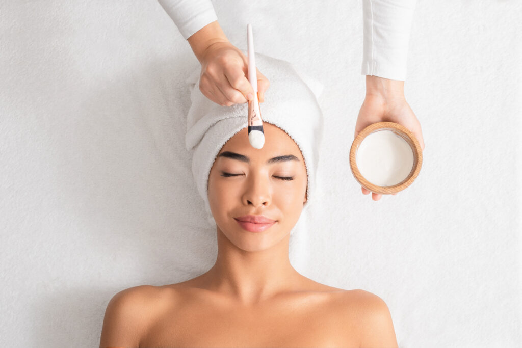 Beauty Treatments. Cosmetologist applying clay mask on asian woman face at spa salon soin esthétique sur le visage